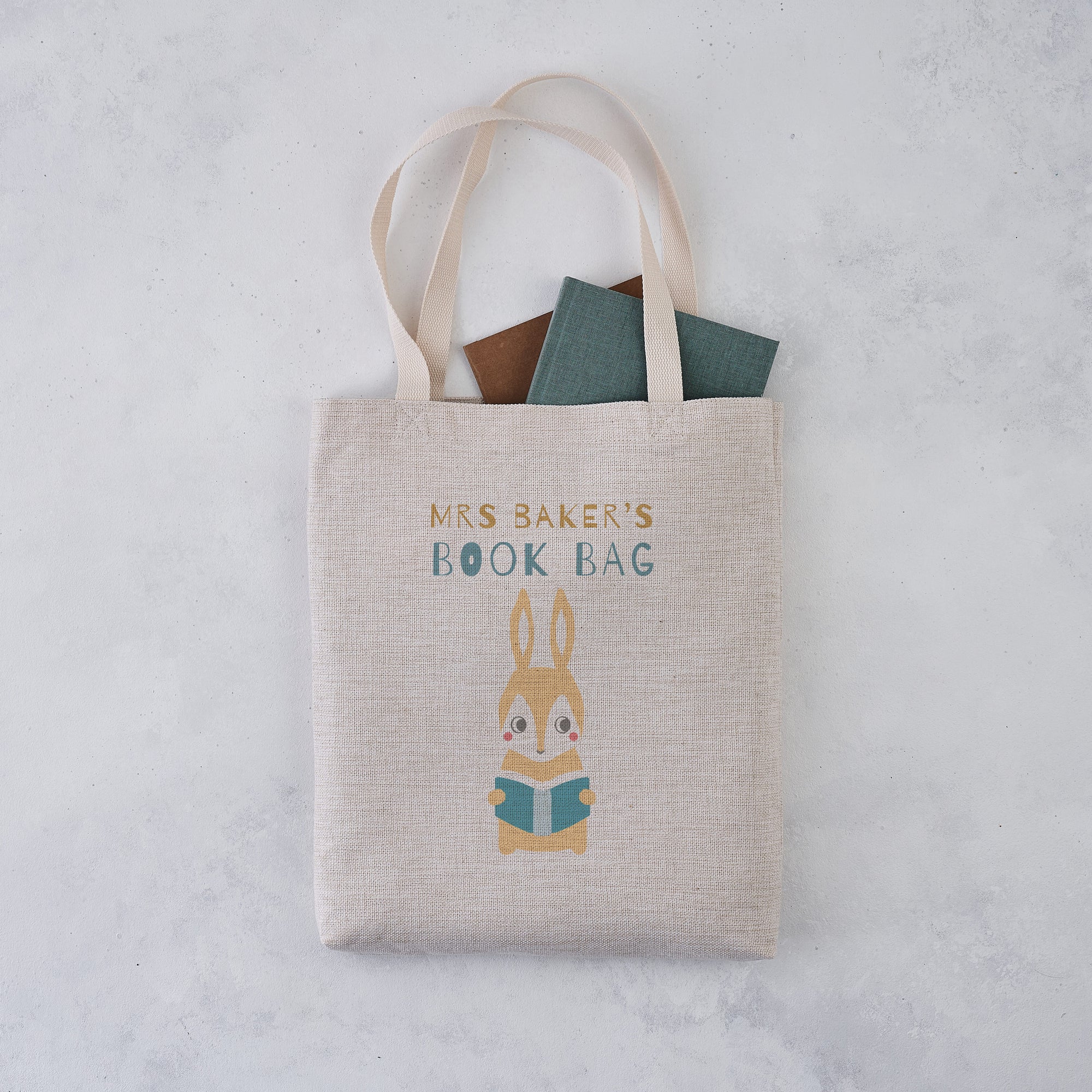 Personalised book bag tote with a rabbit reading a blue book and the text “Mrs Baker’s Book Bag” printed above.