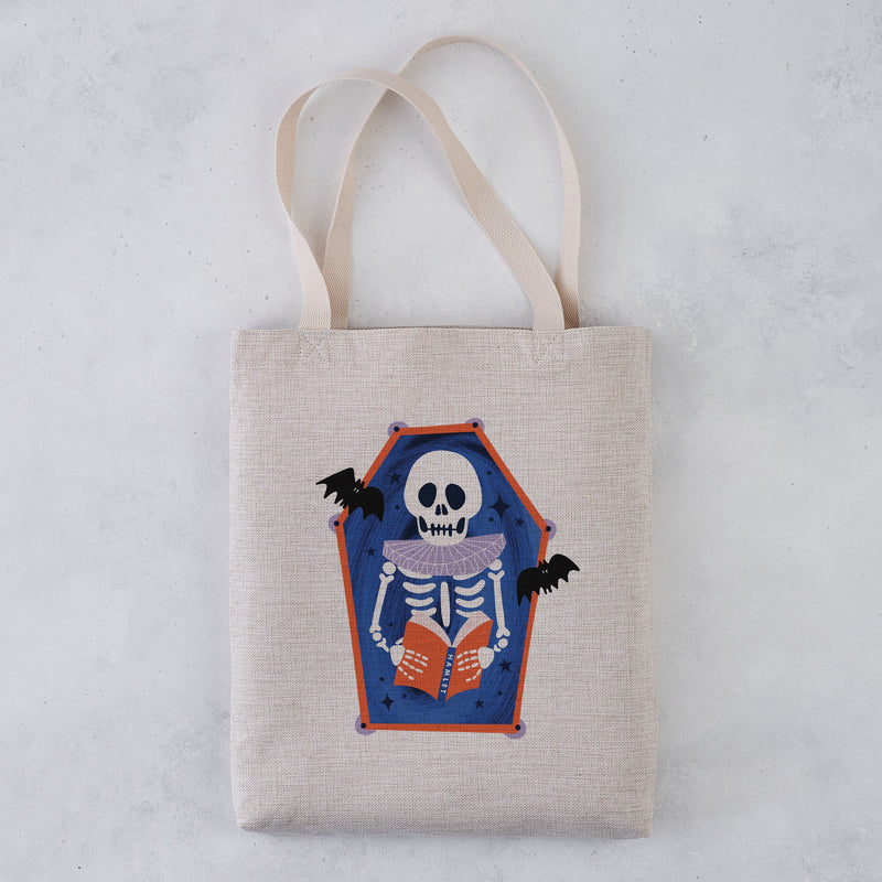 A light beige canvas tote bag with two shoulder straps, featuring a printed coffin-shaped graphic of a skeleton reading an orange book with bats on a blue background, set against a light gray textured backdrop.