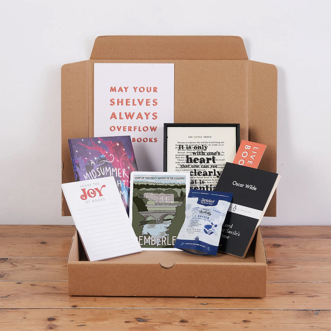 A variety of books and a framed print arranged neatly inside an open brown cardboard crate, placed on a wooden floor.