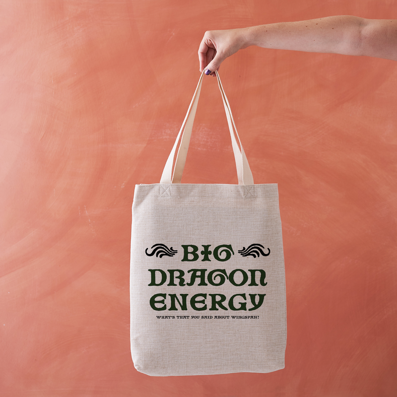 A hand holding a natural canvas tote bag with dark green text that reads 'BIG DRAGON ENERGY' and a smaller tagline underneath, set against a textured peach-pink background.