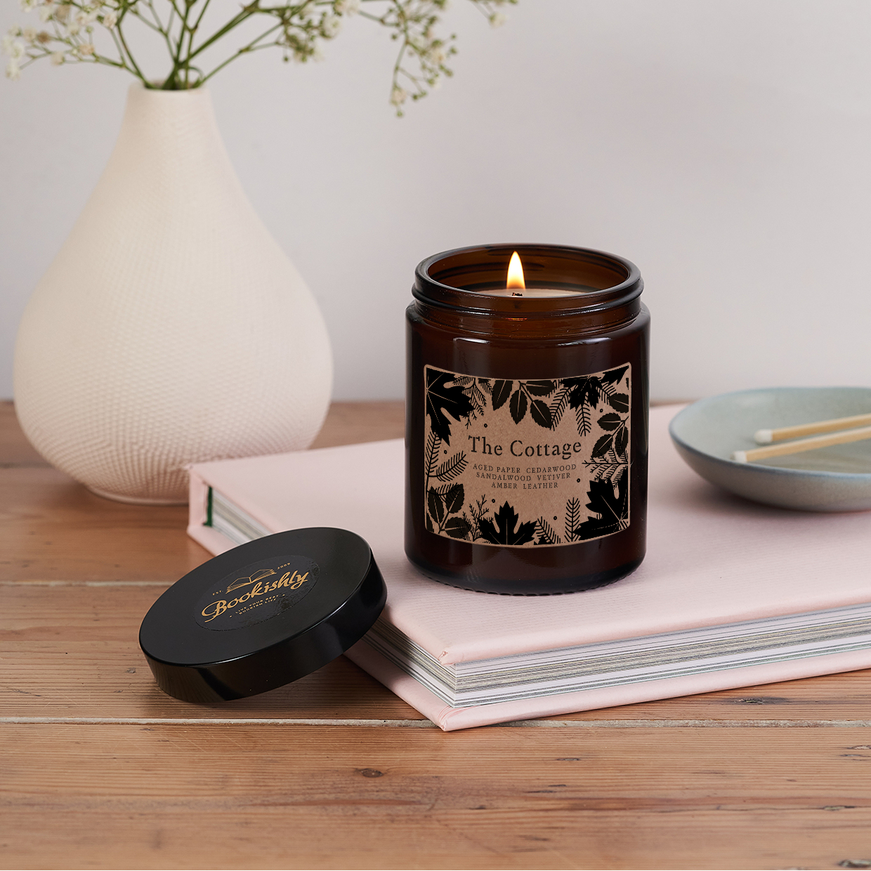Candle in a decorative jar with a floral label on a wooden surface, next to a vase with flowers and a small dish.