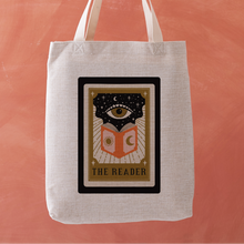 Linen-effect tote bag with tarot card design reading “The Reader,” held against coral background.