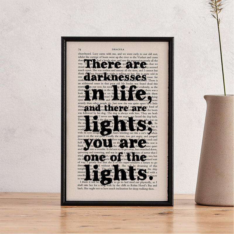 Framed book page print with the quote “There are darknesses in life, and there are lights; you are one of the lights” from Dracula, styled with a tall vase on a wooden table.

