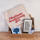 A gift box set with a tote bag, a framed book page print, a packet of luxury tea pyramids, and a book titled 'Persuasion' by Jane Austen, all displayed on a wooden surface.