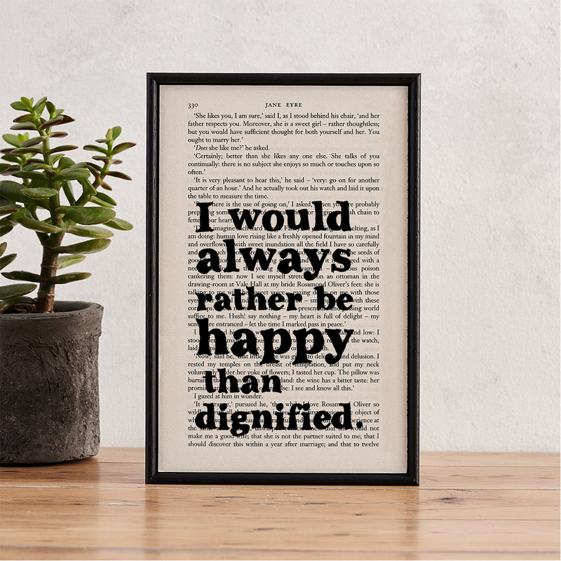 Framed book page print with the quote “I would always rather be happy than dignified” from Jane Eyre, styled with a potted plant on a wooden surface.

