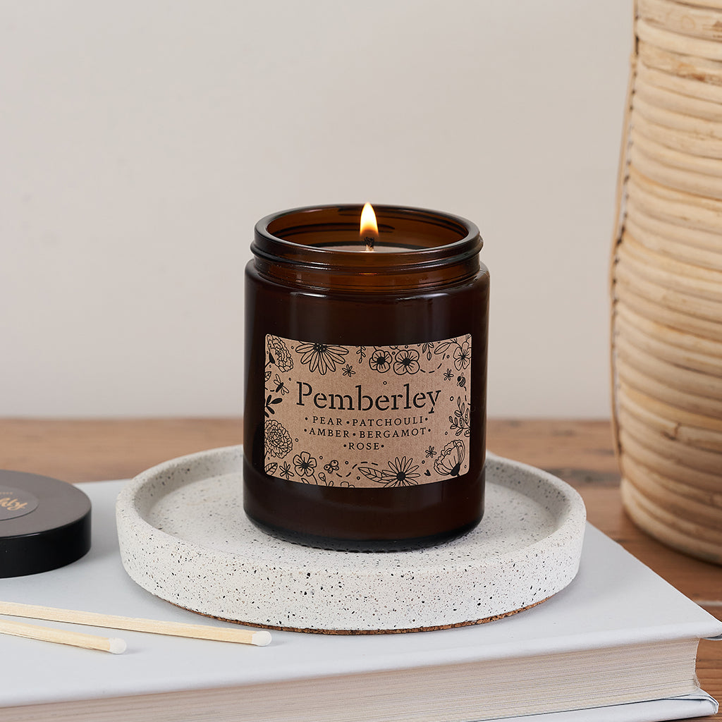 Pemberley candle in amber glass jar with floral kraft label, lit and resting on a speckled ceramic dish beside a Bookishly lid and matches