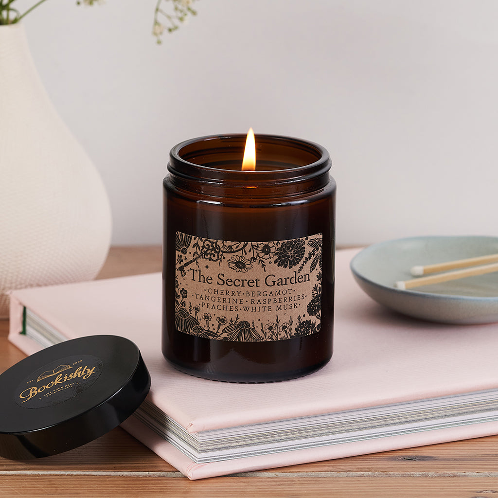 The Secret Garden candle in a dark amber glass jar with floral kraft label, lit and resting on a pale pink book beside a Bookishly lid and ceramic match dish