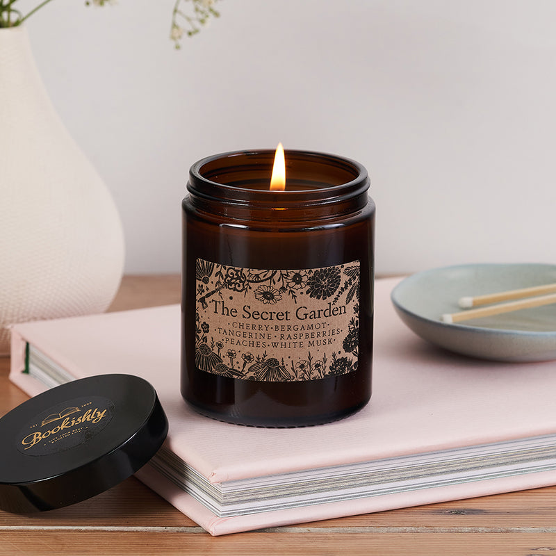 The Secret Garden candle in a dark amber glass jar with floral kraft label, lit and resting on a pale pink book beside a Bookishly lid and ceramic match dish