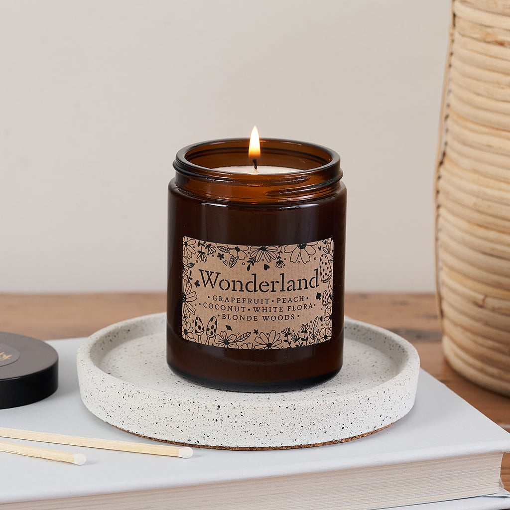 Wonderland candle in amber glass jar with whimsical floral label, lit and resting on a speckled ceramic tray beside matches and a Bookishly lid