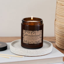 Wonderland candle in amber glass jar with whimsical floral label, lit and resting on a speckled ceramic tray beside matches and a Bookishly lid